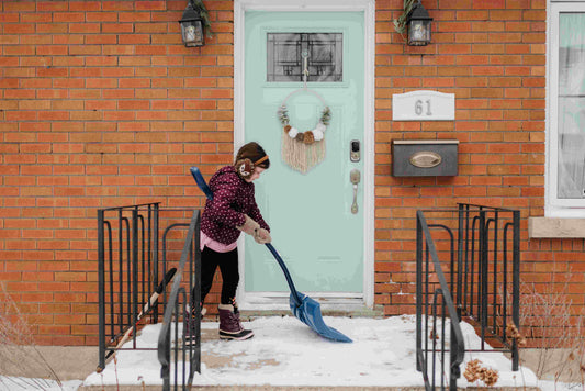 Child shoveling snow in front of a winter home, with a mint-colored door and festive wreath